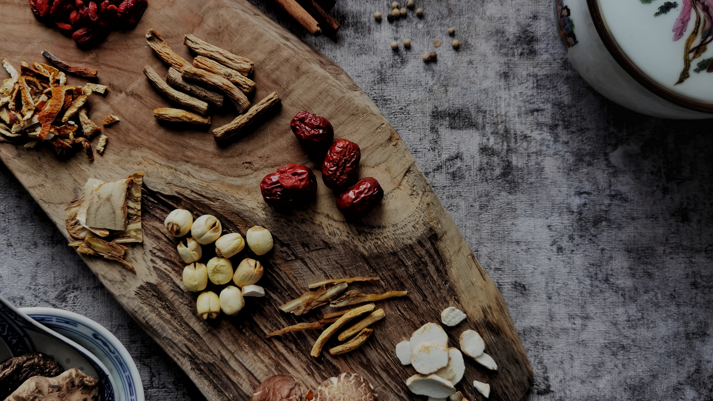 A rustic wooden board displays various dried herbs and ingredients, including red berries, white seeds and sliced roots.